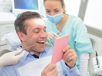 A man is seated in a dental chair, holding a pink card with a surprised expression, while a woman in scrubs stands behind him, smiling and looking at the card.