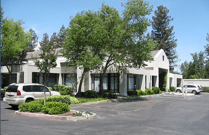 The image depicts a multi-story building with a flat roof, featuring a white exterior and large windows. In front of the building is a parking lot with several cars parked, and there are trees providing shade in the foreground.