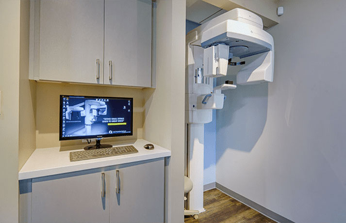 An image of a medical office with a computer monitor and a digital X-ray machine in the foreground, showcasing a modern healthcare environment.