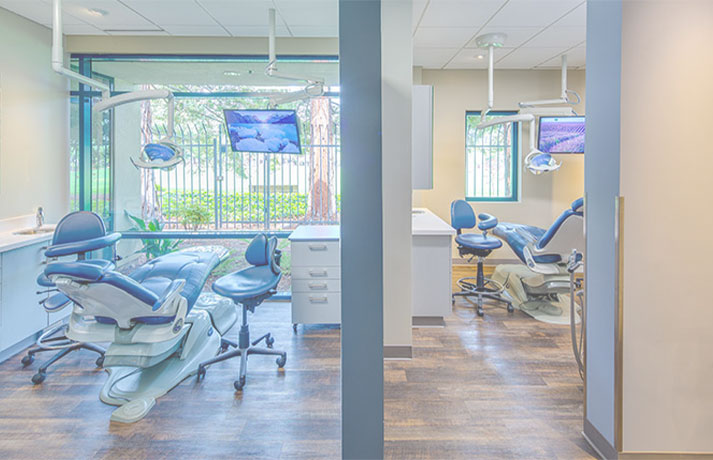 The image shows a modern dental office with two dental chairs, one in the foreground and another in the background. There is a large window allowing natural light to enter, and a television mounted on the wall above the chair in the back.