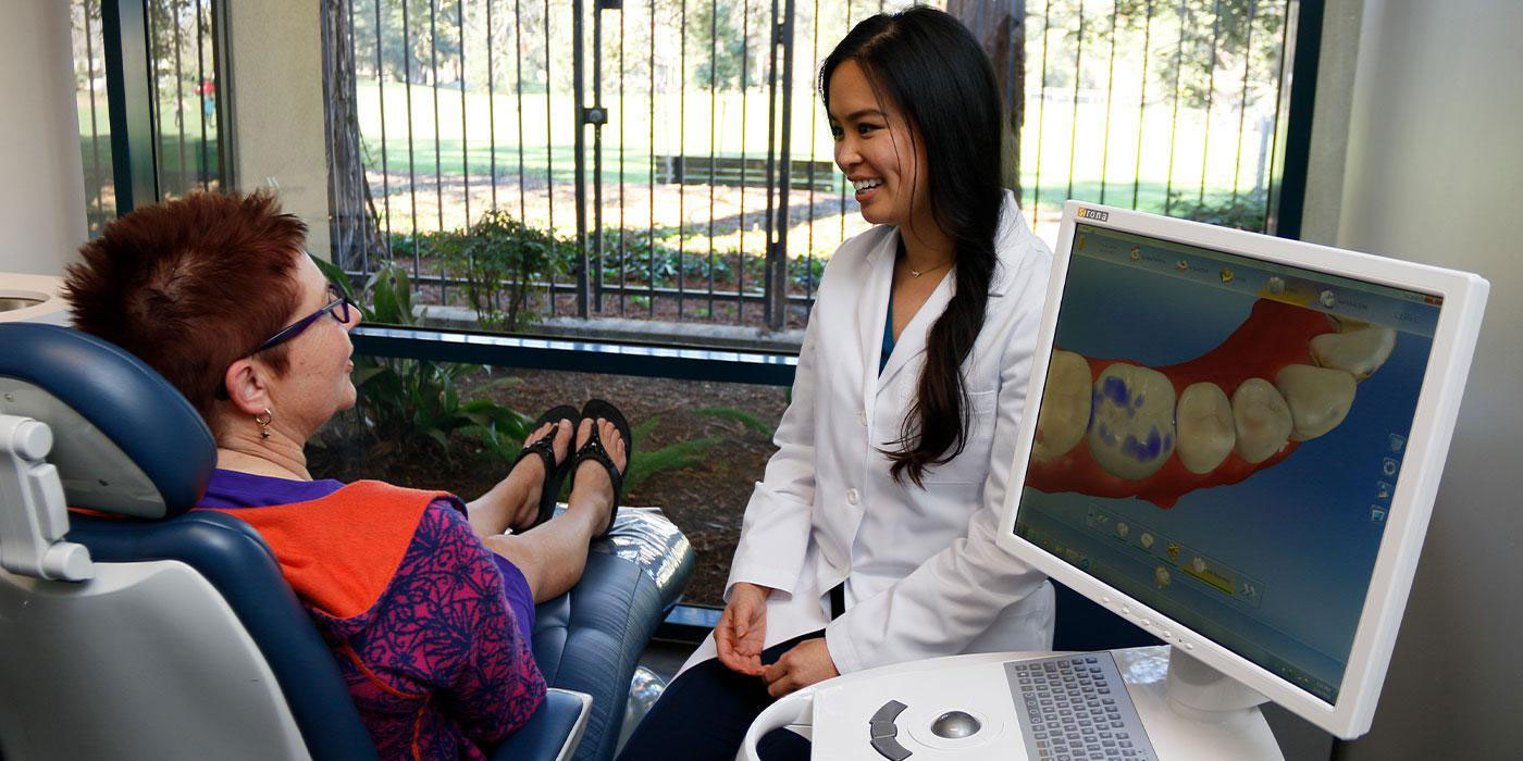 The image shows a dental professional assisting a patient in a dental chair, with the professional using a computer screen and keyboard.
