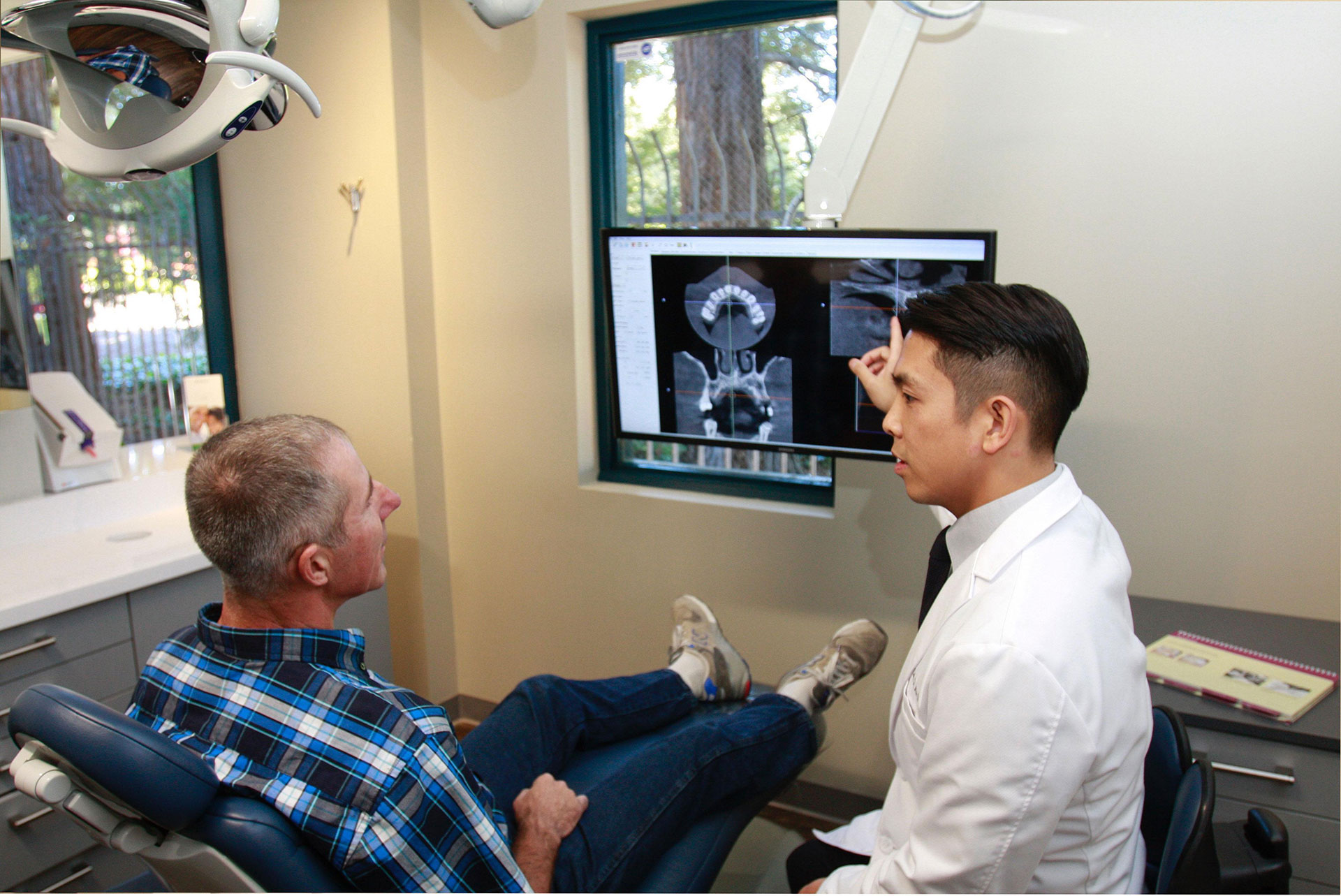 A man is sitting in a dental chair, receiving a check-up from a dentist who is standing next to him. The dentist is pointing at an image of the patient s teeth on a screen behind them.