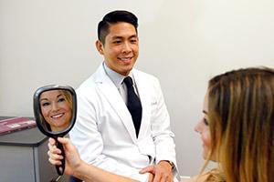 In the image, a man in a lab coat is sitting at a desk with a woman who appears to be a patient. The man has a microphone and is holding up a mirror, seemingly engaged in a conversation or examination with the woman.