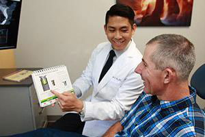 A man in a white lab coat is presenting information to another man, who is seated and holding a pamphlet.