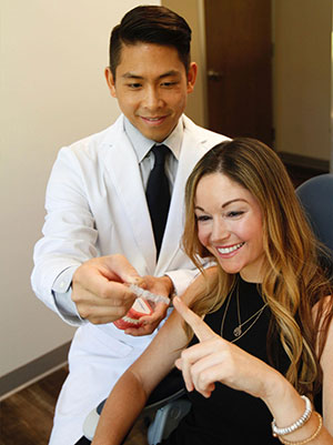 The image depicts a man in a white lab coat holding up a small device, likely a medical or diagnostic tool, to a woman who is pointing at it with her finger.