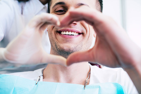 This is a photograph of a man in a dental office, holding up his hands to form a heart shape with his fingers. He appears to be smiling and is wearing a white dental mask.