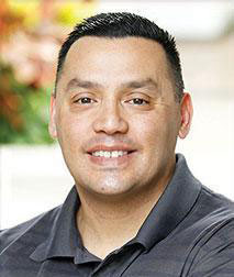 The image is a color photograph featuring an adult male with a beard, smiling and looking directly at the camera. He has short dark hair and appears to be wearing a dark-colored shirt. There are no visible texts or logos in the image that provide additional context about the individual or the setting.