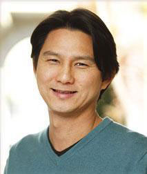 The image is a portrait of an adult male with short hair, wearing a dark top. He has a slight smile and appears to be in a professional setting.