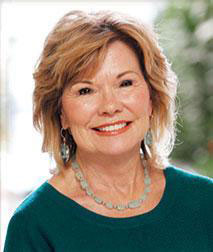 The image is a portrait of an older woman with short hair, wearing a dark top and a necklace. She has a warm smile and is looking directly at the camera.