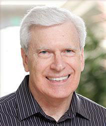 The image shows a smiling man with gray hair, wearing glasses and a dark shirt. He has a beard and mustache, and is standing against a plain background.