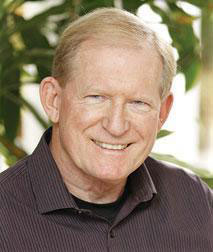 The image features a man with gray hair, wearing glasses and a dark shirt, smiling slightly at the camera. He is standing against a backdrop of greenery.