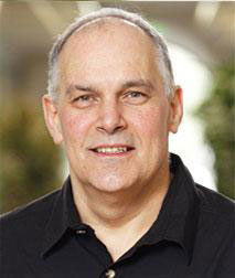 The image is a portrait of an older man with gray hair, wearing a dark shirt, smiling slightly and looking directly at the camera.