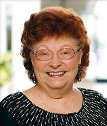 The image is a portrait of an elderly woman with red hair, wearing glasses and a black top, smiling slightly at the camera.