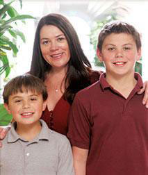 The image is a photograph of three individuals, likely a family, posing together for the camera. A woman stands in the center with two young boys on either side of her; they all appear to be indoors with natural light illuminating the scene.