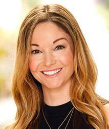 The image is a portrait of a woman with long hair, smiling at the camera. She has light skin and appears to be indoors with natural lighting.