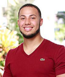 The image is a portrait of a man with a beard and mustache, wearing a red shirt and standing outdoors.