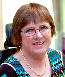 The image is a photograph of an older woman with glasses, smiling at the camera. She has short hair and is wearing a colorful top. The background is blurred but appears to be an indoor setting with natural light.