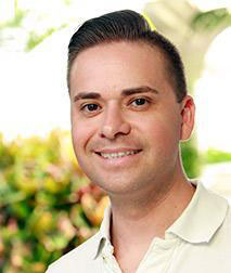 The image shows a man with short hair, wearing a white shirt and smiling at the camera.