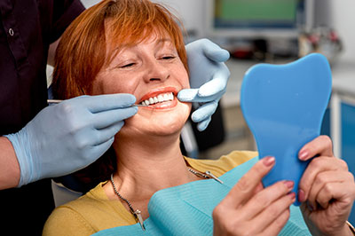 The image shows a woman sitting in a dental chair, smiling broadly, with a dental hygienist performing a cleaning procedure on her teeth.