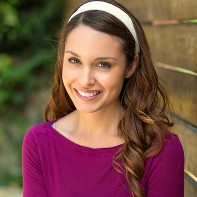 The image shows a woman with long hair smiling at the camera, wearing a purple top and a headband. She is standing against a wooden fence in an outdoor setting.