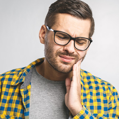 The image shows a man with glasses, wearing a blue plaid shirt and holding his hand to his mouth as if he s in pain or discomfort.
