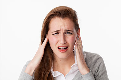 A young woman with blonde hair, wearing a gray sweater and holding her head with both hands, expressing concern or pain.